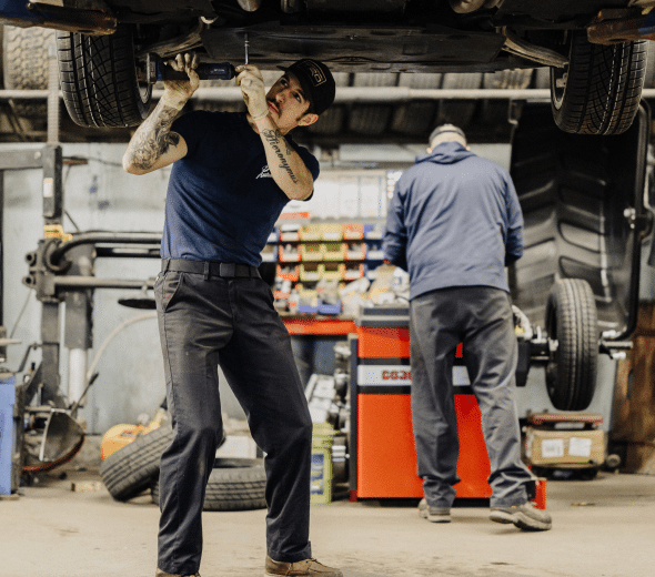 Two mechanics working in a garage; one inspecting a vehicle's underside, the other organizing tools near a workbench.