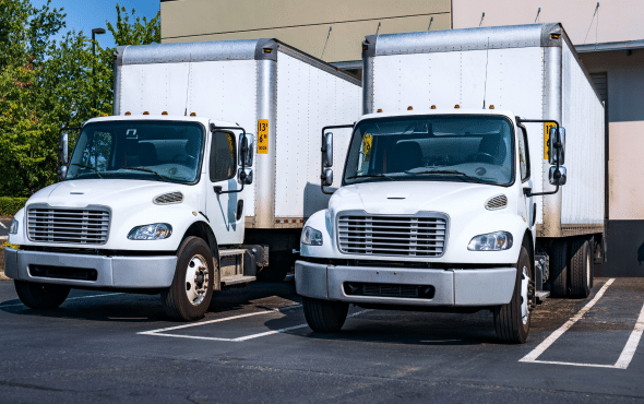 Two white box trucks parked side by side in a parking lot under a clear sky.