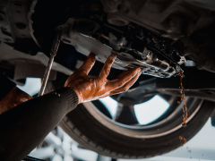 Mechanic's hands working on car engine underside with oil dripping, wheel visible in the background.