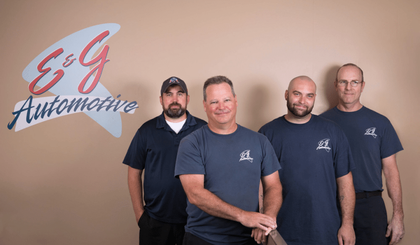 Four men in blue shirts pose in front of an "E&G Automotive" sign on a beige wall.