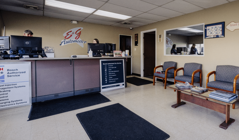 A waiting area with four chairs, a reception desk, and two people working behind the counter. Various brochures are on a table.