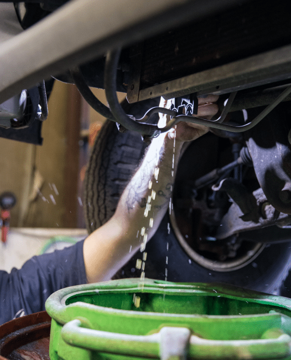 A person's hand draining car oil into a green container from the undercarriage of a vehicle in a garage.