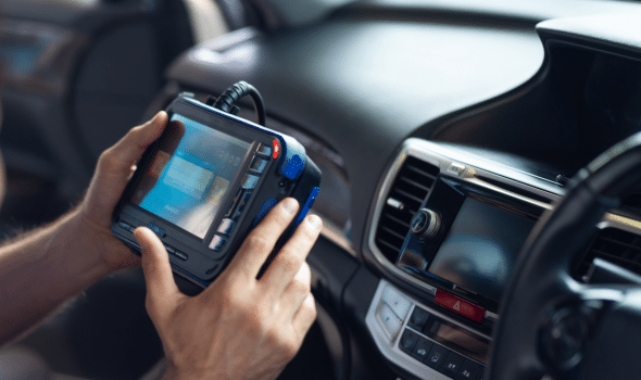 A person using a diagnostic tool in a car, holding the device near the dashboard and steering wheel.