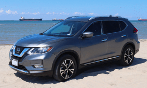 Gray Nissan crossover SUV parked on a sandy beach with ocean and ships in the background.